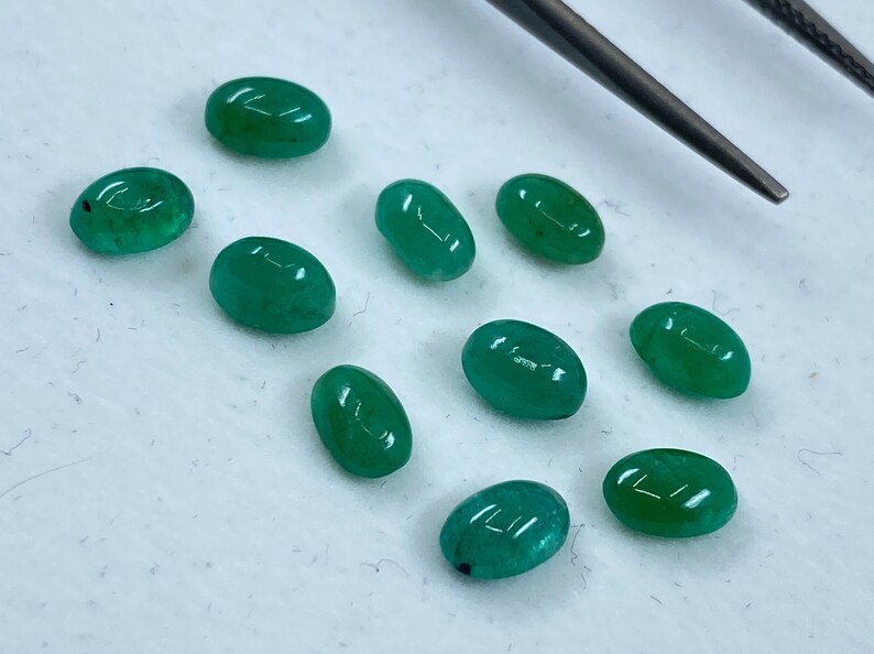 May include: Ten oval-shaped green gemstones, likely emeralds, are arranged on a white surface. A metal tool is visible in the background.
