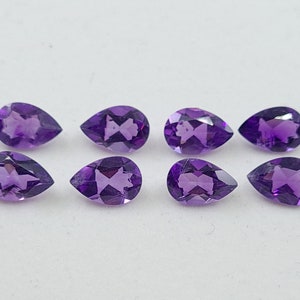 May include: Eight pear-shaped amethyst gemstones, each with a deep purple hue, are arranged in two rows of four on a white surface.