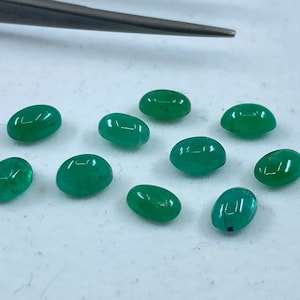 May include: Ten oval-shaped green gemstones, likely emeralds, are scattered on a white surface.