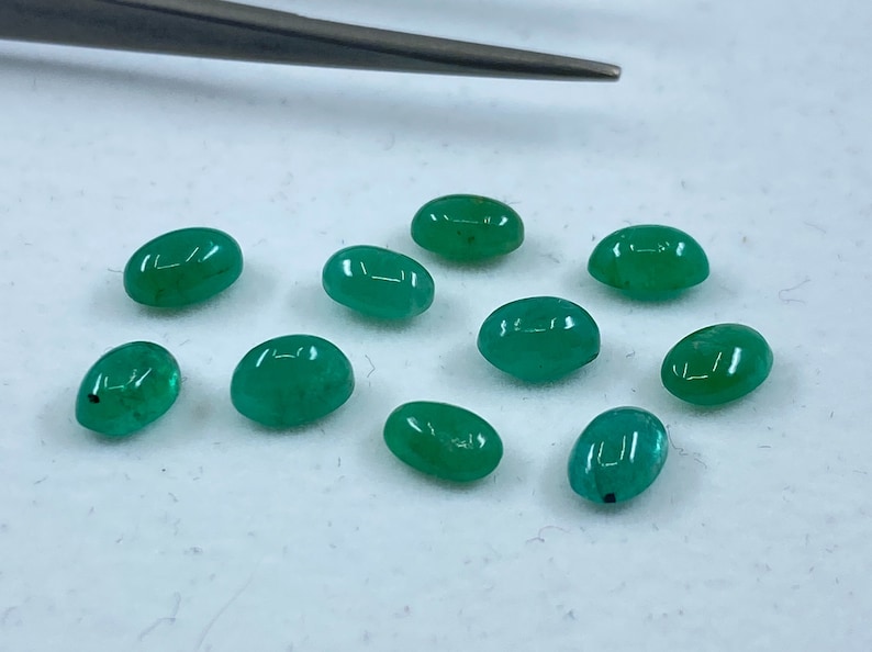May include: Ten oval-shaped green gemstones, likely emeralds, are scattered on a white surface.