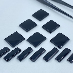May include: A collection of black gemstone cabochons in square and rectangular shapes. The cabochons are polished and have a smooth, glossy finish.