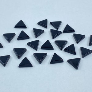 Puede incluir: Una colección de 20 gemas de vidrio de forma triangular negra. Las gemas están esparcidas sobre una superficie blanca.