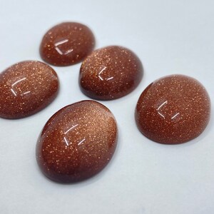 May include: Five oval-shaped, polished gemstones with a sparkling, glittery appearance. The stones are a warm, reddish-brown color and have a smooth, domed surface. They are arranged on a white background.