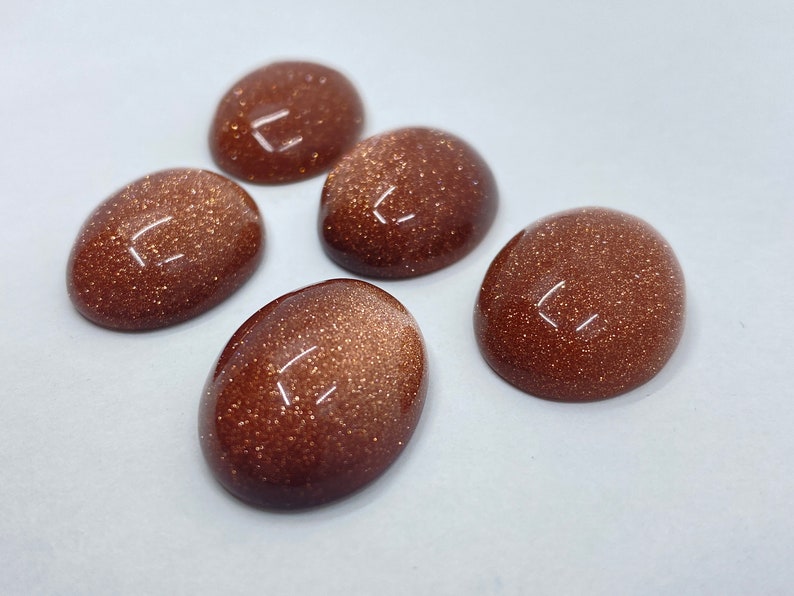 May include: Five oval-shaped, polished gemstones with a sparkling, glittery appearance. The stones are a warm, reddish-brown color and have a smooth, domed surface. They are arranged on a white background.