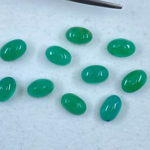 May include: Ten oval-shaped green gemstones, likely emeralds, are arranged on a white surface. The gemstones are smooth and polished, with a slight variation in size and shade.