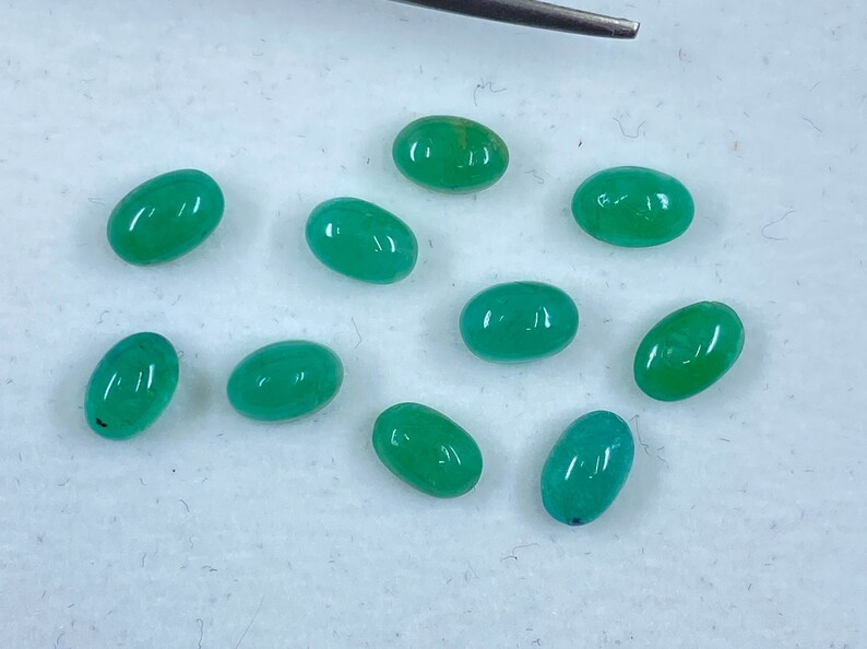 May include: Ten oval-shaped green gemstones, likely emeralds, are arranged on a white surface. The gemstones are smooth and polished, with a slight variation in size and shade.