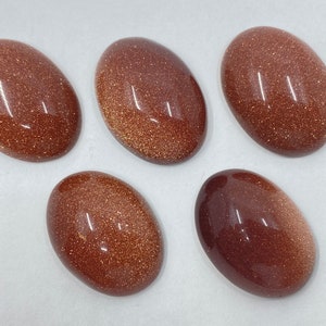 May include: Five oval-shaped, polished gemstones with a sparkling, golden-brown appearance. The stones have a smooth, reflective surface and vary slightly in color, ranging from a deep brown to a lighter, shimmering gold. These stones are ideal for jewelry making.