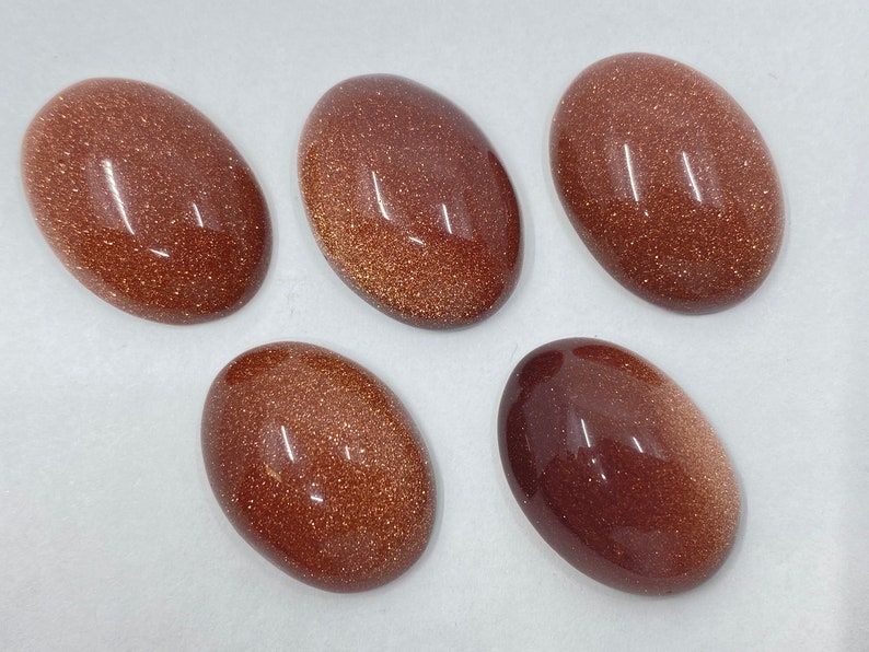 May include: Five oval-shaped, polished gemstones with a sparkling, golden-brown appearance. The stones have a smooth, reflective surface and vary slightly in color, ranging from a deep brown to a lighter, shimmering gold. These stones are ideal for jewelry making.