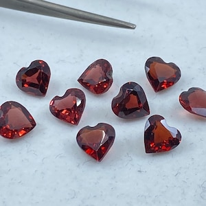 Puede incluir: Ocho piedras preciosas rojas en forma de corazón, probablemente granates, están dispuestas sobre una superficie blanca. Las piedras preciosas están facetadas y tienen un acabado brillante.