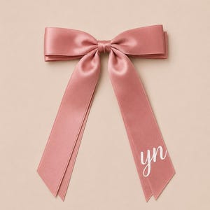 Peut inclure: Un nœud pour cheveux en satin rose poudré avec une finition lisse et brillante. Le nœud possède deux longs pans tombants, avec les lettres minuscules "yn" imprimées en blanc dans une police cursive sur le bas du pan droit de l'accessoire.