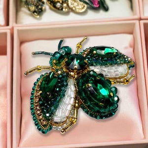 May include: A decorative insect-shaped brooch featuring deep emerald green faceted crystals, clear white glass beads, and gold-toned metal accents. The design incorporates intricate beadwork and metallic wire detailing to form the wings and body.