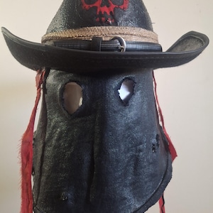 May include: A black hat with a wide brim and a red skull graphic on the front. The hat features a tan woven band with a black buckle strap. A black fabric face covering with two eye holes and frayed red fabric strips hangs from the hat brim.