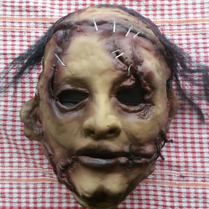 May include: A realistic horror mask with stitched-up wounds and a sewn-shut mouth. The mask is made of latex and has a realistic skin tone. It features black hair and a distressed look.