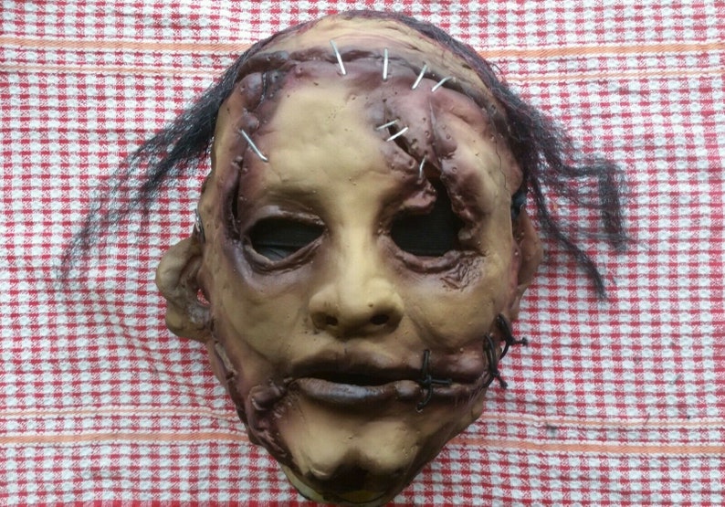 May include: A realistic horror mask with stitched-up wounds and a sewn-shut mouth. The mask is made of latex and has a realistic skin tone. It features black hair and a distressed look.