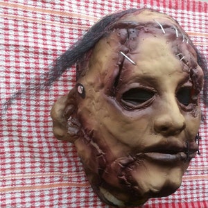 May include: A realistic horror mask with stitched-up wounds and a distressed, flesh-toned appearance. The mask has black hair and a stitched-up mouth.