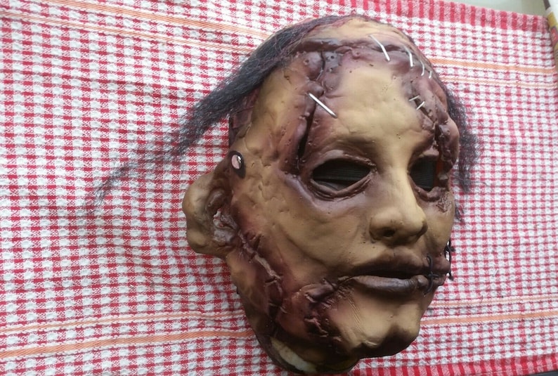 May include: A realistic horror mask with stitched-up wounds and a distressed, flesh-toned appearance. The mask has black hair and a stitched-up mouth.