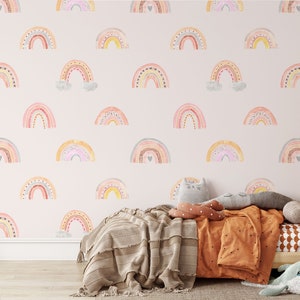 Puede incluir: Una pared de guarder&iacute;a rosa claro con un patr&oacute;n repetitivo de arco&iacute;ris de acuarela coloridos. Los arco&iacute;ris est&aacute;n decorados con peque&ntilde;os puntos y corazones. La pared est&aacute; detr&aacute;s de una cama de madera con una manta y almohadas marrones.