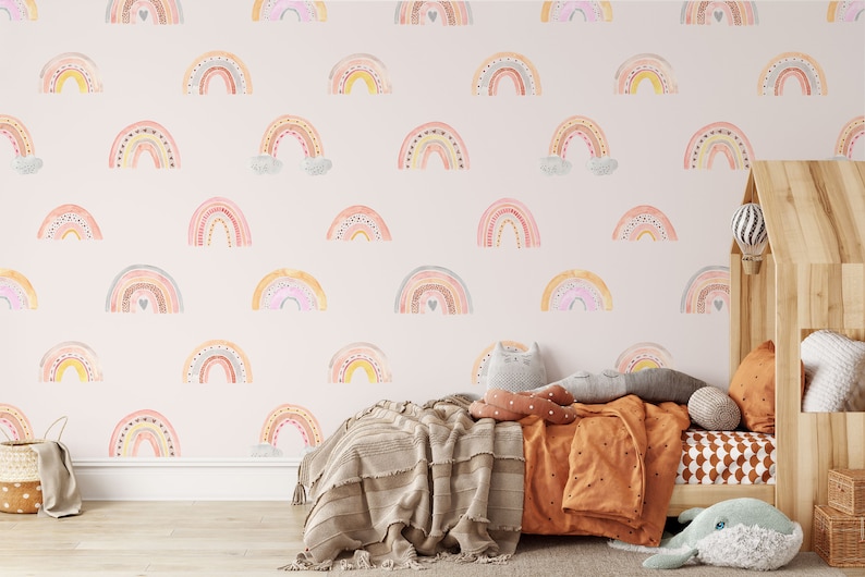 Puede incluir: Una pared de guarder&iacute;a rosa claro con un patr&oacute;n repetitivo de arco&iacute;ris de acuarela coloridos. Los arco&iacute;ris est&aacute;n decorados con peque&ntilde;os puntos y corazones. La pared est&aacute; detr&aacute;s de una cama de madera con una manta y almohadas marrones.