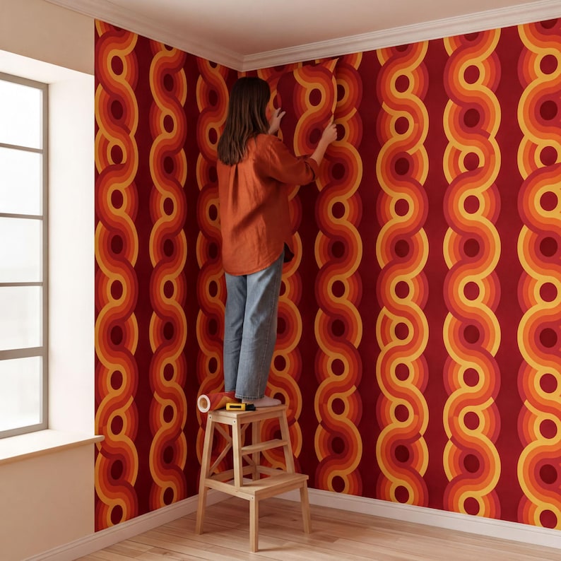 A person is standing on a stool and applying wallpaper to a wall with a vibrant, retro-style pattern in shades of red, orange, and yellow.