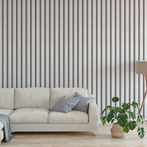 Puede incluir: Un sofá seccional blanco con dos almohadas de color azul y una manta gris se encuentra frente a una pared con paneles de madera verticales. Una lámpara de pie con una pantalla beige y una planta verde en maceta se encuentran a la derecha de la imagen.
