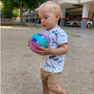 May include: A young child in a white t-shirt with a colorful animal print holds a pink, blue, and green ball with a cartoon pig on it.