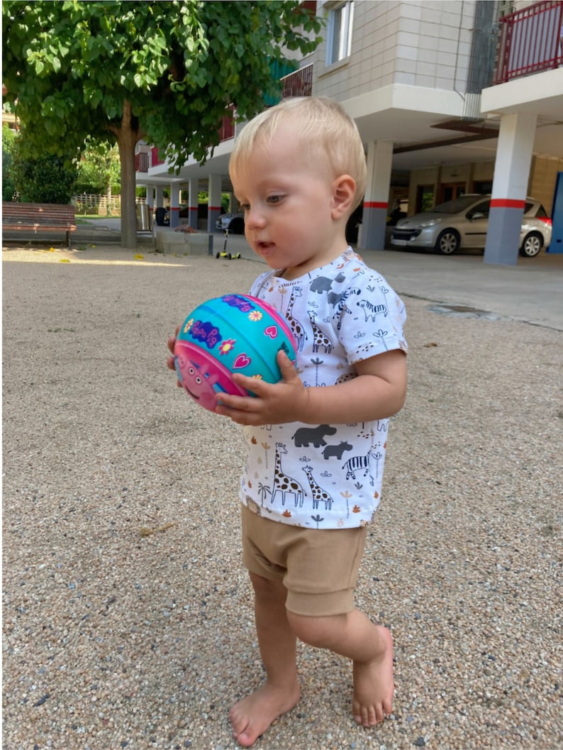 May include: A young child in a white t-shirt with a colorful animal print holds a pink, blue, and green ball with a cartoon pig on it.