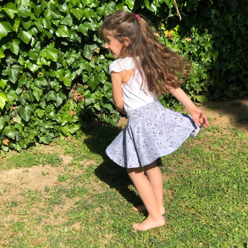 May include: A white short-sleeved top with flutter sleeves paired with a light grey flared skirt featuring a repeating pattern of small cartoon characters, stars, and circular motifs, worn in a spinning motion outdoors.