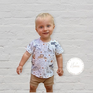 May include: A toddler wearing a white short-sleeved t-shirt with a brown, black, and white animal print. The shirt has a round neckline and short sleeves. The toddler is also wearing brown shorts. The image includes a logo for "Handsome Pattern" with the text "Letter A4 AOPattern PDF & Tutorial Newborn - 3 Years".