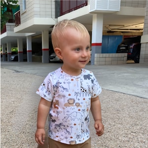 May include: A young child wearing a white short-sleeved t-shirt with a colorful print of animals and plants. The shirt has a crew neck and a relaxed fit.
