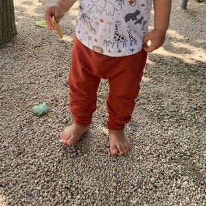 Op de afbeelding: Een wit shirt met korte mouwen voorzien van een herhalend patroon van bruine en zwarte safaridieren, waaronder giraffen, olifanten en zebra's, gecombineerd met een effen roestkleurige broek met een relaxte pasvorm en boorden bij de enkels.