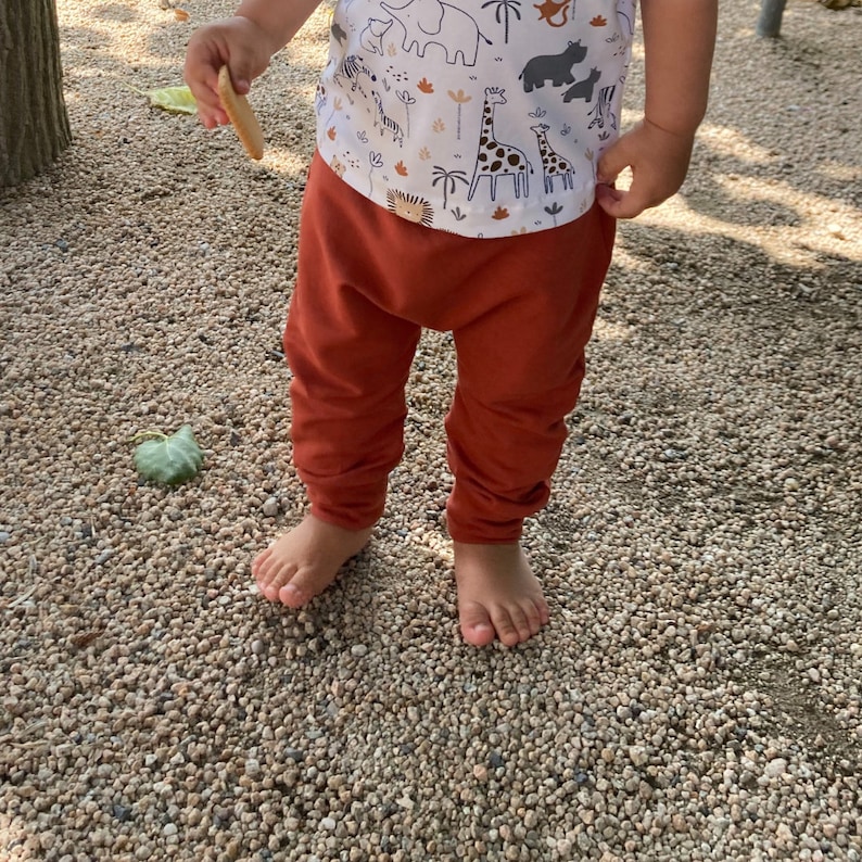 Op de afbeelding: Een wit shirt met korte mouwen voorzien van een herhalend patroon van bruine en zwarte safaridieren, waaronder giraffen, olifanten en zebra's, gecombineerd met een effen roestkleurige broek met een relaxte pasvorm en boorden bij de enkels.