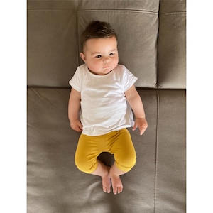 Puede incluir: Un beb&eacute; de cabello oscuro viste una camiseta blanca lisa de manga corta y unos leggings de color amarillo mostaza. El infante est&aacute; acostado boca arriba, luciendo un conjunto sencillo compuesto por una prenda superior clara y pantalones amarillos.
