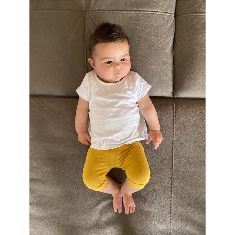 Puede incluir: Un beb&eacute; de cabello oscuro viste una camiseta blanca lisa de manga corta y unos leggings de color amarillo mostaza. El infante est&aacute; acostado boca arriba, luciendo un conjunto sencillo compuesto por una prenda superior clara y pantalones amarillos.