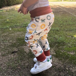 Op de afbeelding: Off-white joggingbroek met een herhalend patroon van veelkleurige X- en O-symbolen in blauw-, oranje- en geeltinten. De broek heeft een brede, effen roestkleurige elastische tailleband en bijpassende geribbelde enkelboorden.