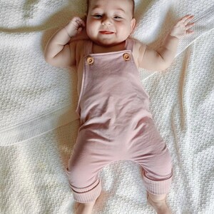 May include: A baby in a pale pink jumpsuit with wooden button fastenings on the straps. The jumpsuit features ribbed cuffs at the ankles. The baby is lying on a white textured blanket, smiling and reaching out with both arms.