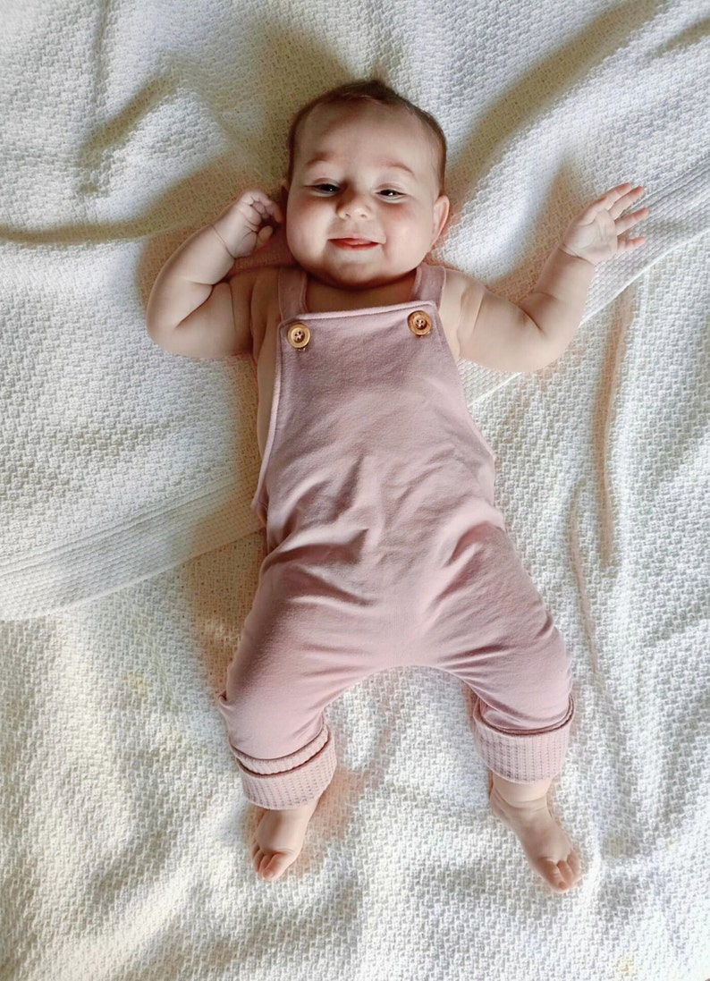 May include: A baby in a pale pink jumpsuit with wooden button fastenings on the straps. The jumpsuit features ribbed cuffs at the ankles. The baby is lying on a white textured blanket, smiling and reaching out with both arms.