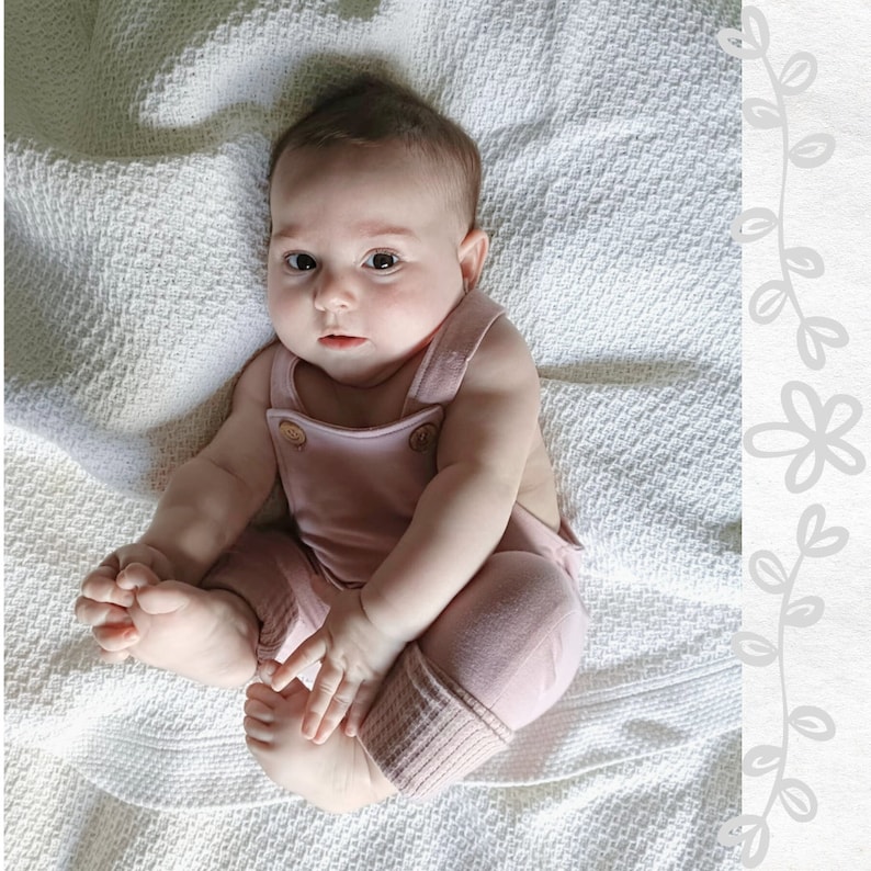 May include: A baby wearing dusty pink overalls with ribbed cuffs and small metallic buttons at the chest straps. The garment features a simple design with wide shoulder straps and a soft fabric texture, providing a comfortable fit for an infant.