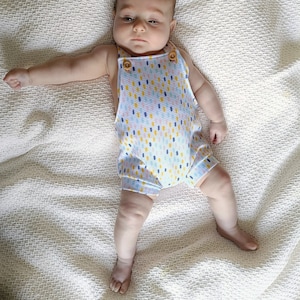 May include: A sleeveless romper featuring a white base with a repeating pattern of small, vertical dashes in yellow, blue, and orange. The garment has two light brown wooden buttons at the top of the front straps and a textured, waffle-like fabric.