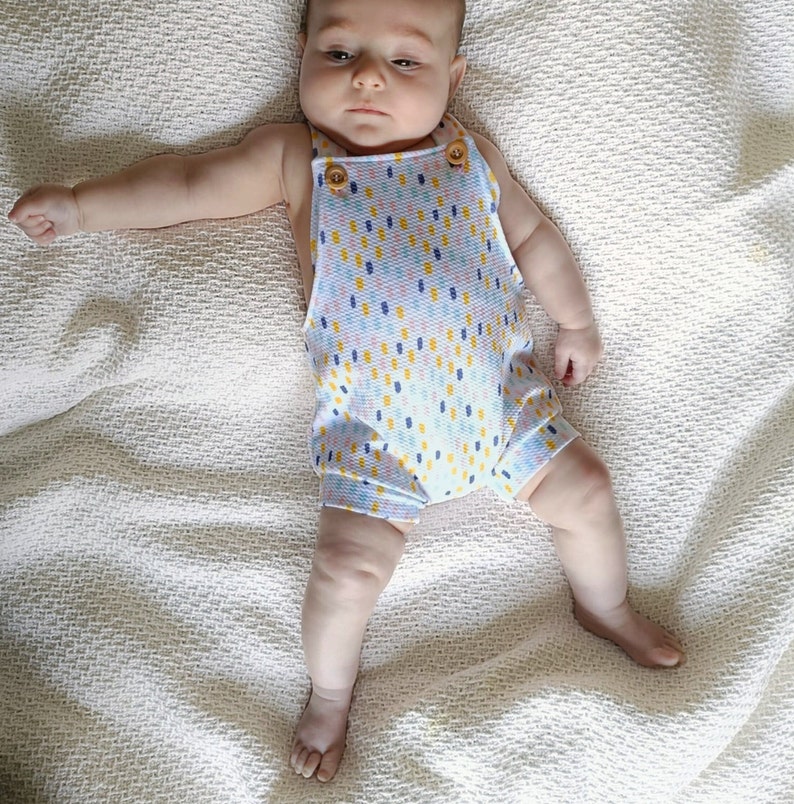 May include: A sleeveless romper featuring a white base with a repeating pattern of small, vertical dashes in yellow, blue, and orange. The garment has two light brown wooden buttons at the top of the front straps and a textured, waffle-like fabric.