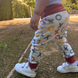 Op de afbeelding: Lichtgrijze broek met een herhalend patroon van handgetekende X- en O-symbolen in geel, blauw en rood. De broek heeft een brede, effen donkerrode tailleband en bijpassende donkerrode enkelboorden, gecombineerd met witte sneakers met klittenband.
