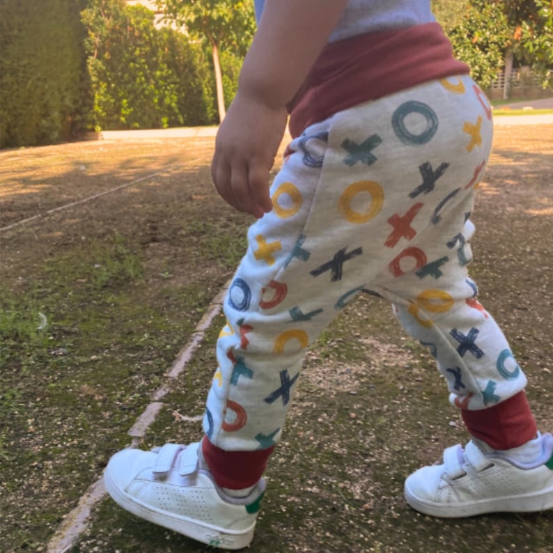 Op de afbeelding: Lichtgrijze broek met een herhalend patroon van handgetekende X- en O-symbolen in geel, blauw en rood. De broek heeft een brede, effen donkerrode tailleband en bijpassende donkerrode enkelboorden, gecombineerd met witte sneakers met klittenband.