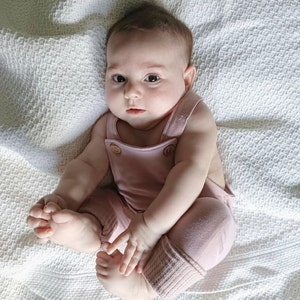 May include: A baby in a dusty rose romper suit with wooden buttons and ribbed cuffs. The baby is sitting on a white textured blanket. The romper has suspender straps. The baby is looking at the camera.