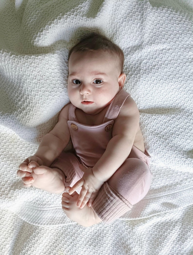 May include: A baby in a dusty rose romper suit with wooden buttons and ribbed cuffs. The baby is sitting on a white textured blanket. The romper has suspender straps. The baby is looking at the camera.