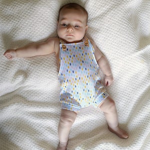 May include: A baby in a light blue romper with a colourful dotted pattern. The romper features wooden button fastenings on the straps. The baby is lying on a white textured blanket.