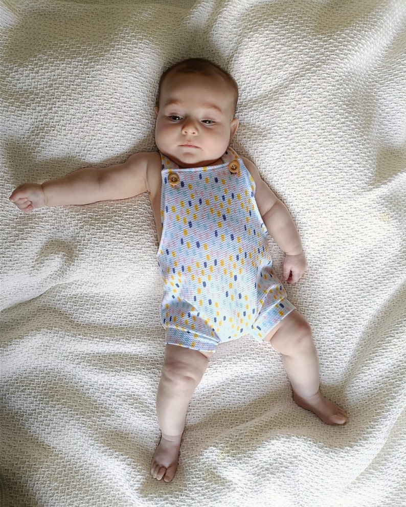 May include: A baby in a light blue romper with a colourful dotted pattern. The romper features wooden button fastenings on the straps. The baby is lying on a white textured blanket.