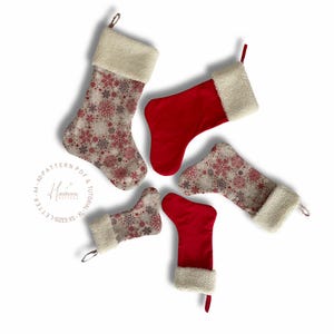 Peut inclure: Cinq bas de Noël disposés en étoile. Deux sont rouges unis, et trois sont ornés de flocons de neige rouges et gris sur fond crème. Chaque bas a un revers crème et une boucle pour l'accrocher.