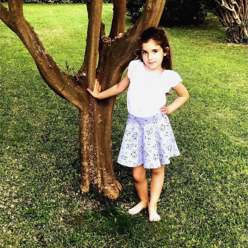 May include: A white short-sleeved top with ruffled sleeves paired with a light purple skirt featuring a repeating black and white pattern of small animal illustrations. The skirt has a wide waistband and a flared silhouette that reaches above the knees.