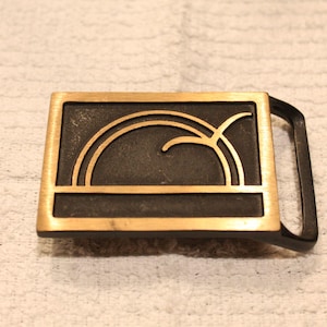 May include: Rectangular belt buckle with a black background and a gold-colored border. The buckle features a gold-colored geometric design. The buckle has a black handle on the right side. The buckle is made of metal.