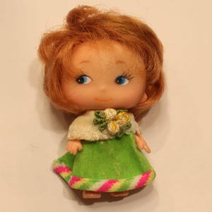 May include: A small doll with short, wavy ginger hair, blue eyes, and a light skin tone. The doll wears a white, textured shawl with small yellow and green floral accents over a bright green dress featuring a striped pink, yellow, and white hem.