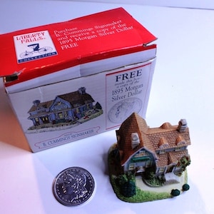 May include: A miniature house model with a brown roof and green trim, sitting on a base with green grass and small trees. The house is part of a Liberty Falls collection and comes with a free replica 1895 Morgan Silver Dollar.
